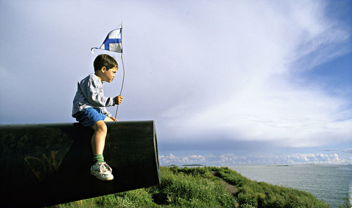 Side view of boy with finnish flag sitting on cannon against sky