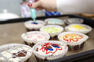 A woman squeezes colored frosting from a tube onto chocolate brown cupcakes covered  white frosting.