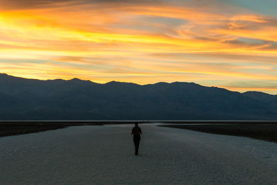 Silhouette man walking dirt road leading towards mountains against orange sky
