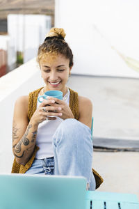 Happy young woman holding tea cup sitting with laptop at table on rooftop