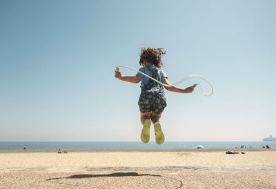 Girl playing with skipping rope while jumping on beach against clear sky