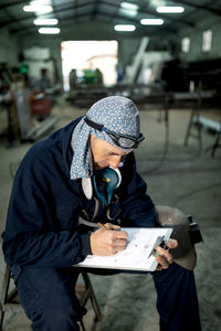 Serious male worker in hardhat and overall taking notes in notebook with pen on table near metal constructions in light garage