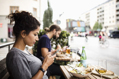 Side view of woman text messaging while having food at sidewalk cafe in city