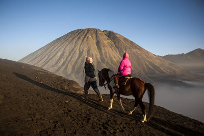Rear view of person riding horse while man walking on mountain against sky