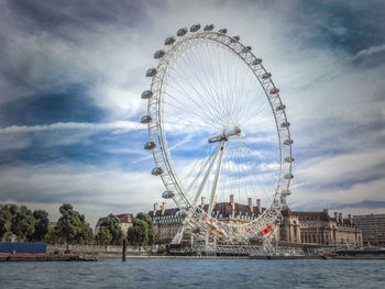 Ferris wheel by river against cloudy sky