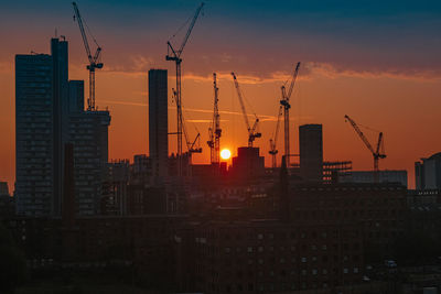 Silhouette cranes by buildings against sky during sunset