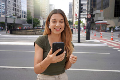 Excited brazilian girl wearing earphones watching her smartphone in city street