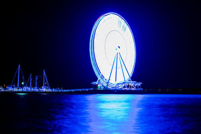 Illuminated ferris wheel by sea against clear sky at night