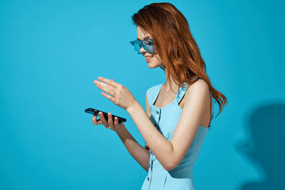 Young woman using phone while standing against blue sky