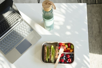 Rye bread, berries and salad with disposable fork in lunch box on table by water bottle and laptop