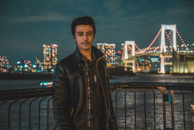 Portrait of young man standing against bridge in city at night