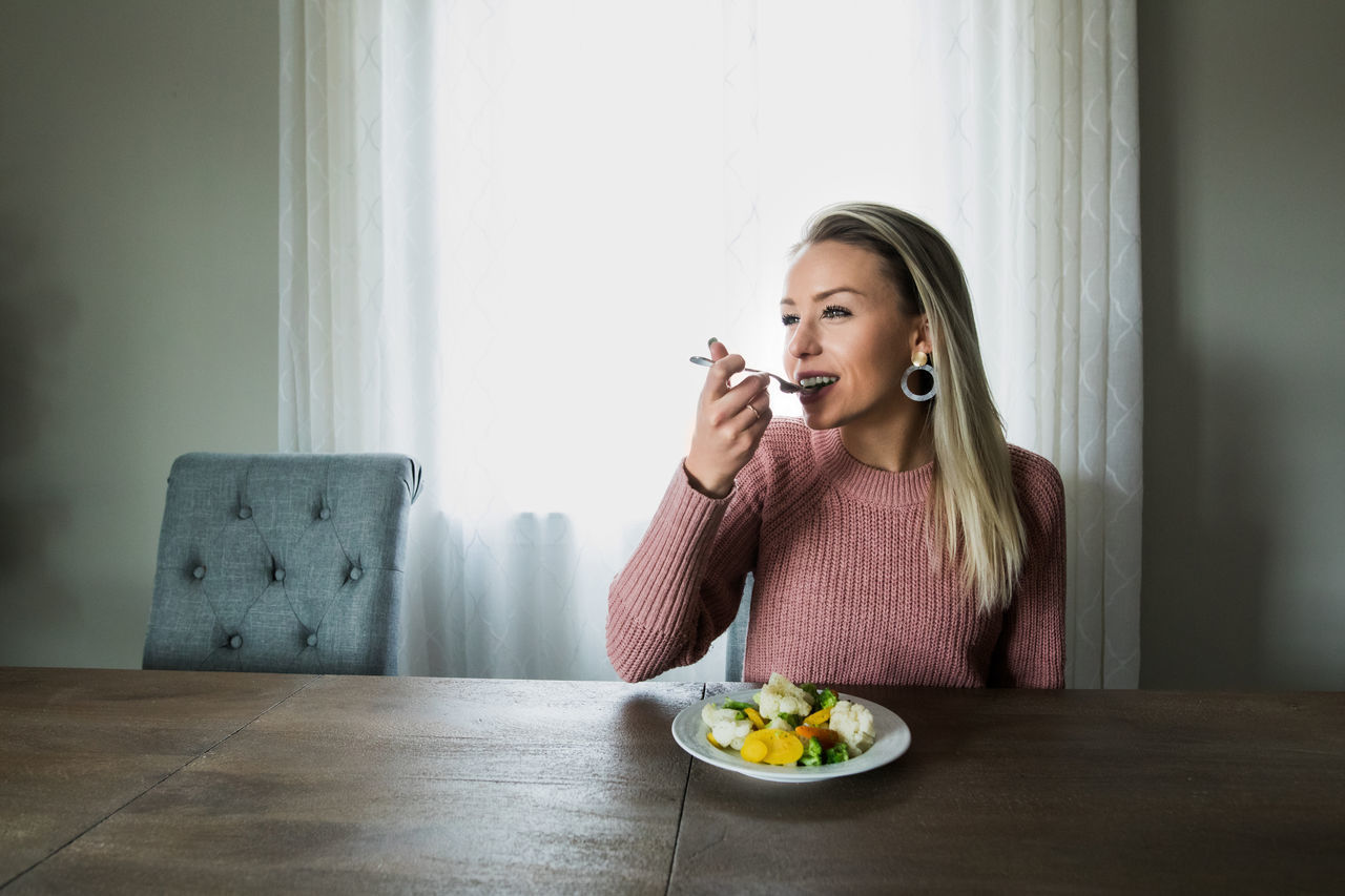 food and drink, one person, food, sitting, indoors, eating, blond hair, young adult, lifestyles, holding, hair, eating utensil, young women, front view, table, casual clothing, healthy eating, wellbeing, hairstyle, meal