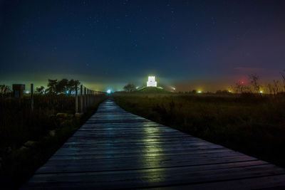 Empty boardwalk against illuminated lights at night