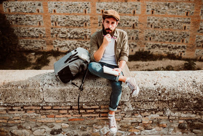 Thoughtful man holding book and pencil while sitting on retaining wall