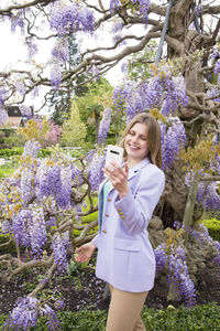 A young woman in a lilac jacket makes selfie with the flowering wisteria