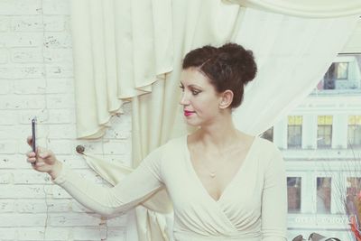 Woman taking selfie while standing against curtain at home