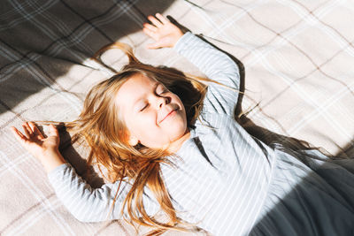 Cuty funny little girl with long hair in grey home dress having fun lies in sun light on bed at home