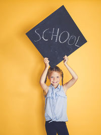 Portrait of smiling girl holding blackboard with school text while standing against yellow background