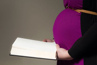 Midsection of pregnant woman holding book against wall