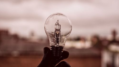 Close-up of hand holding light bulb against sky