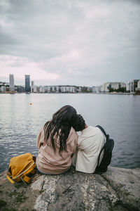 Rear view of teenage boy with head on shoulder of female friend while sitting on rock near sea in city