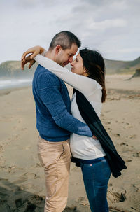 Side view of couple embracing while standing on beach against sky