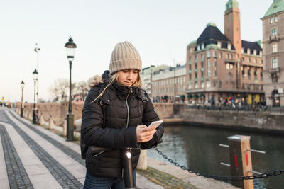 Teenage girl using smart phone while standing on e-scooter by canal in city