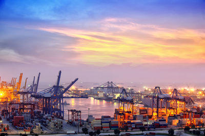 Cranes at harbor against sky during sunset