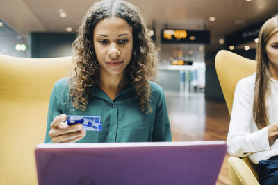 Confident businesswoman holding credit card while using laptop by colleague at airport lobby