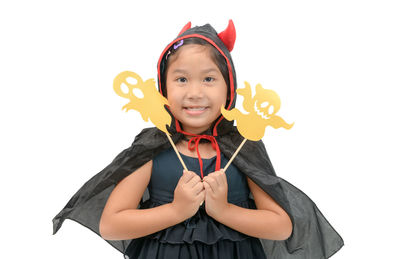 Portrait of smiling girl in spooky costume holding ghost props while standing against white background during halloween