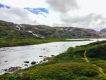 Stream flowing by hills against cloudy sky