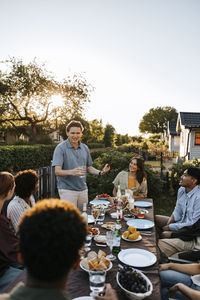 Young man giving speech to multiracial group of male and female friends sitting at dinner party in back yard