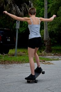 Rear view full length of woman with arms outstretched riding longboard on road