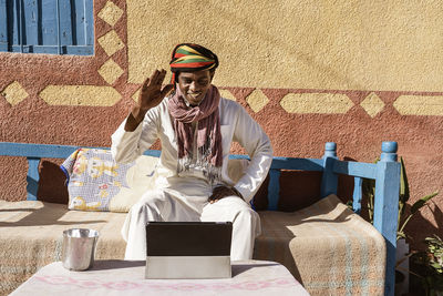 Sincere egyptian male in traditional turban and robe having video chat on tablet while sitting on sofa in backyard on sunny day