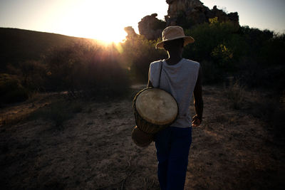 Rear view of man with musical instrument standing against trees during sunset