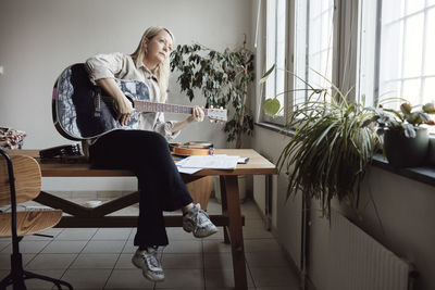 Mature woman playing guitar while sitting on table in classroom