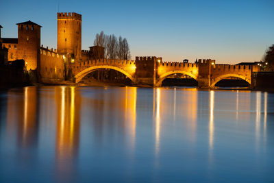 Arch bridge over river by illuminated buildings against sky