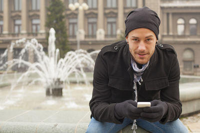 Man using mobile phone while sitting against fountain
