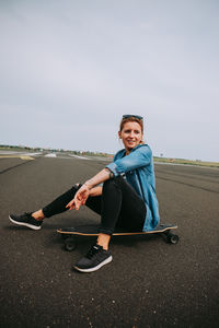 Smiling woman sitting on skateboard against sky