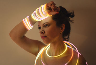 Portrait of woman with arms raised wearing neon bracelet standing against gray background