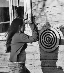 Side view of young woman aiming on dartboard against wooden wall