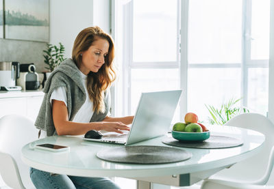 Young adult blonde woman with long hair working on laptop sitting on kitchen at home, remote work