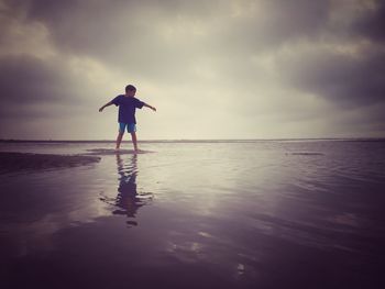 Full length of boy with arms outstretched standing at beach against sky