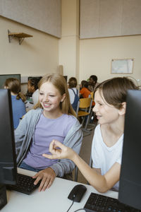 Smiling boy talking to female friend using computer while sitting in classroom at school