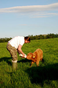 Side view of woman feeding milk to calf on grassy field against sky