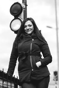 Portrait of young woman with hands in pockets standing by road signal against clear sky