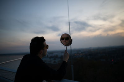 Side view of man holding mirror with his reflection against sky during sunset