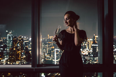 Woman drinking champagne at home against cityscape during night