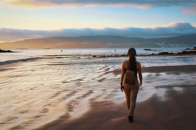 Girl with a backpack walking on a beach at dusk with low tide.