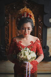 Young woman in traditional clothing holding bouquet while standing against wall